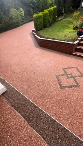 A wide terracotta resin driveway with two interlocking squares inlaid in dark aggregate, edged against a red brick retaining wall